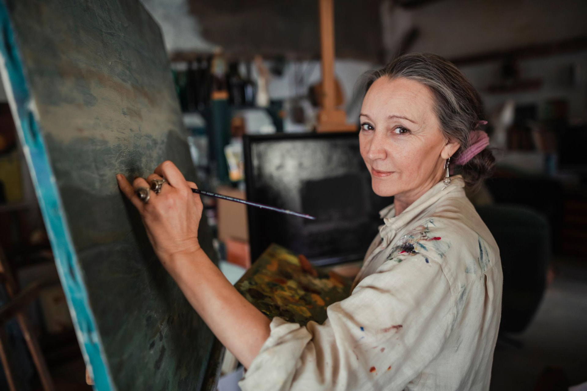 A woman working on a large canvas in her studio, brush in hand, looking up