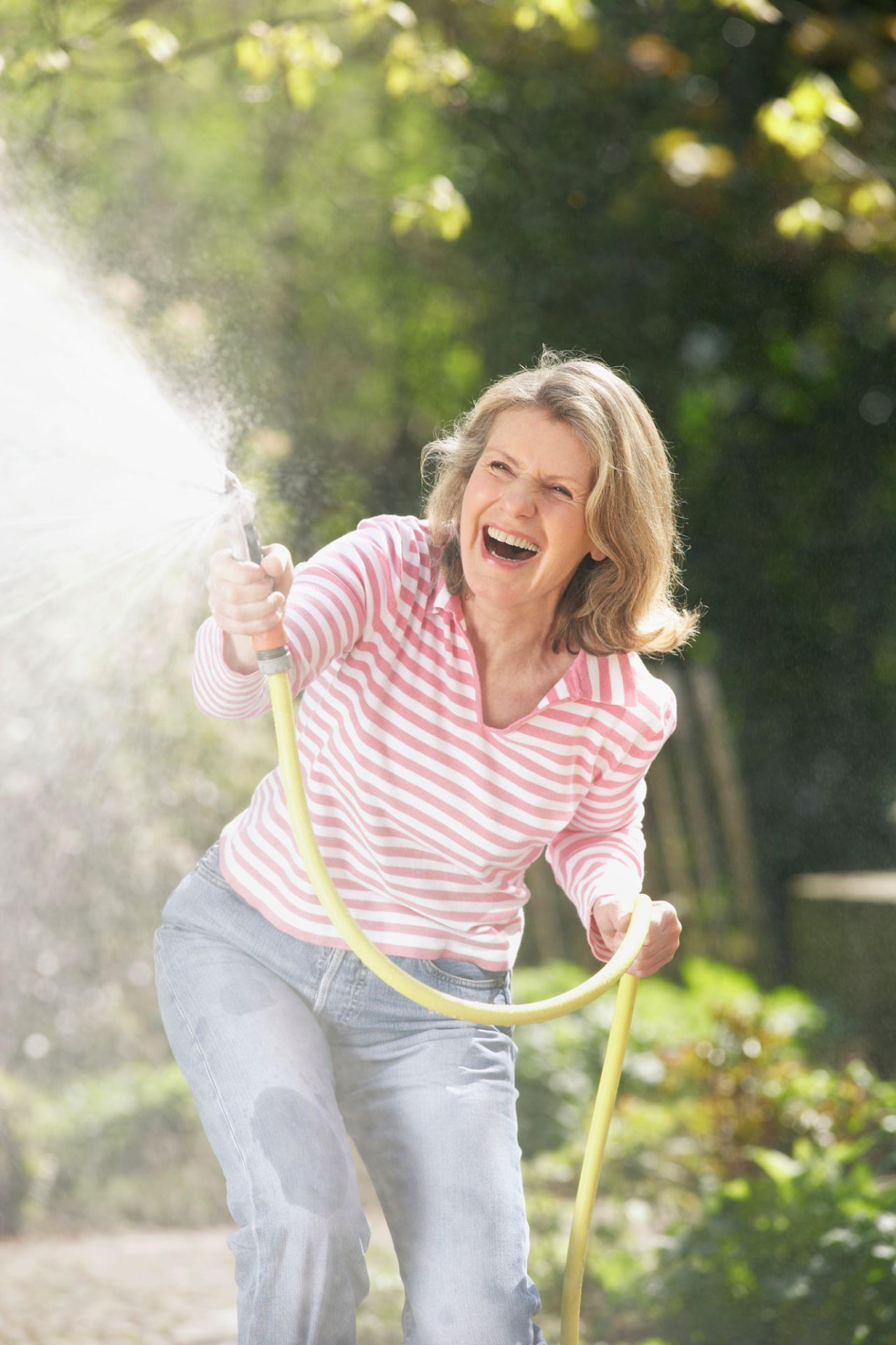 A woman in the second half of life laughing in her garden while spraying a hose