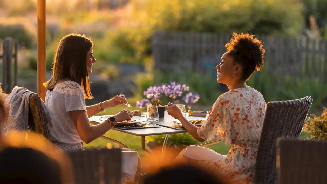 Two women sitting across from each other at an outdoor table in warm evening light, talking over a meal