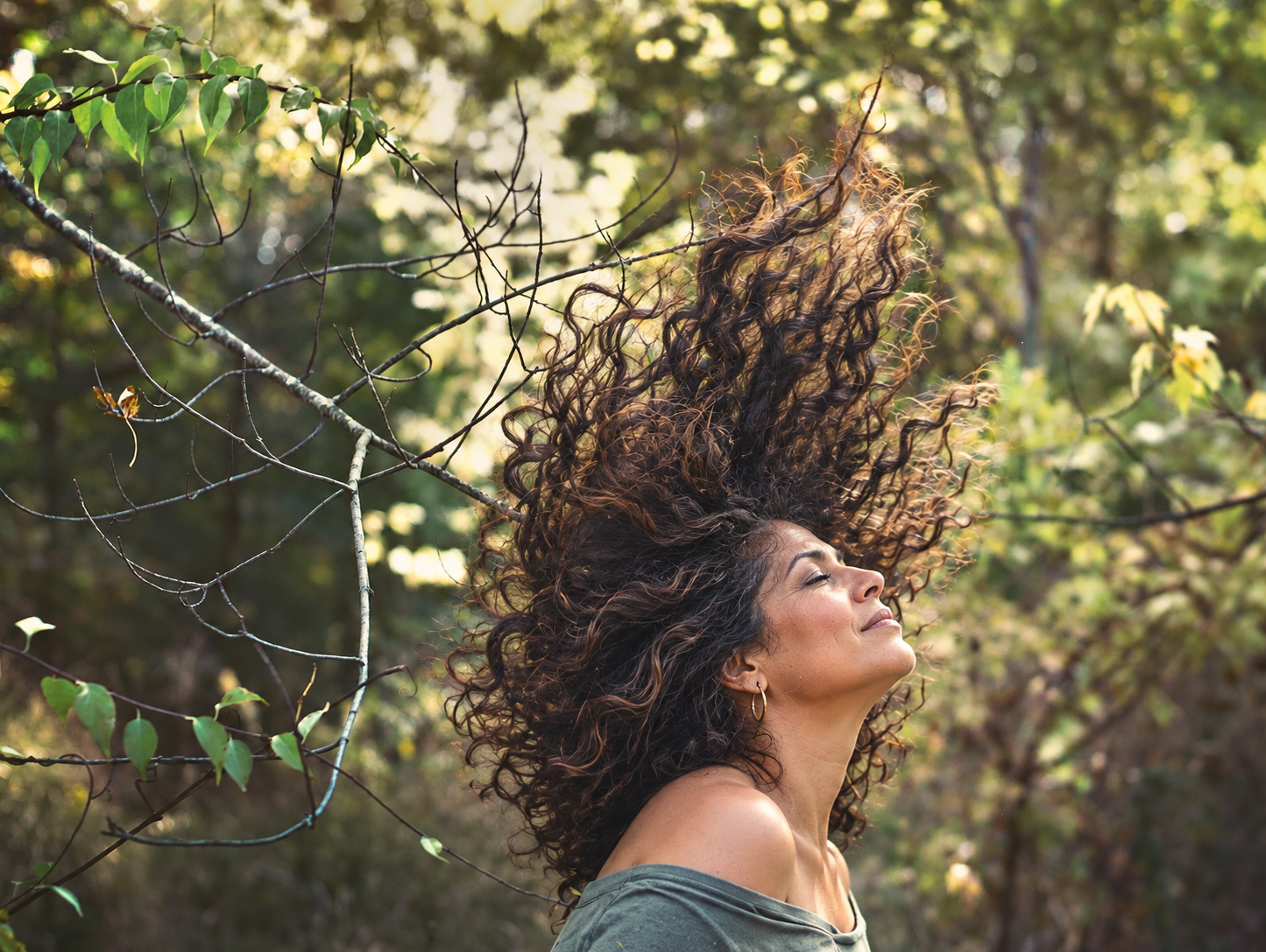 Middle-aged Latina woman with dark curly hair lifted dramatically upward in a sunlit wooded setting.