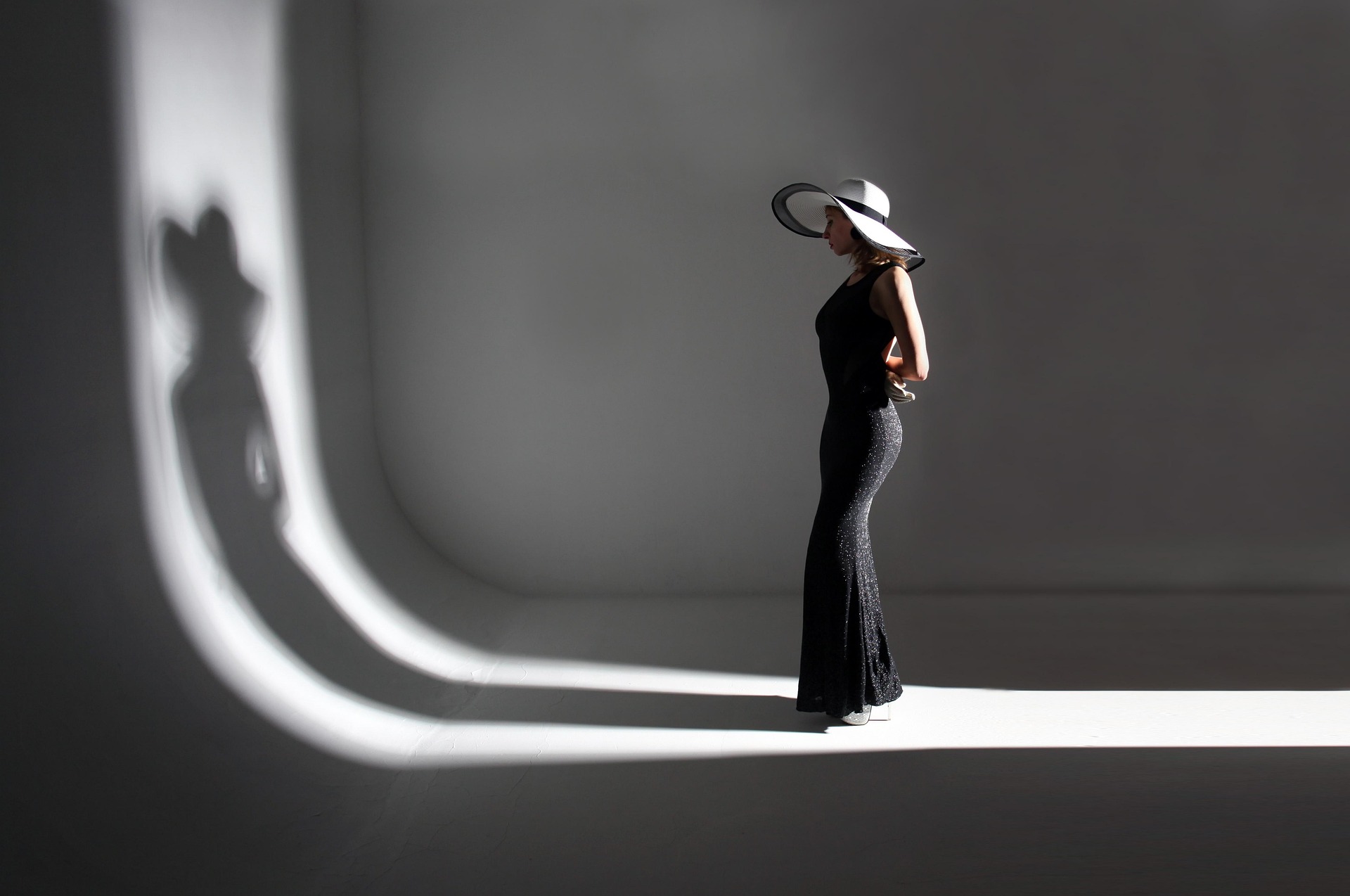 A woman in a wide-brimmed hat standing in a band of light cast by a curved window